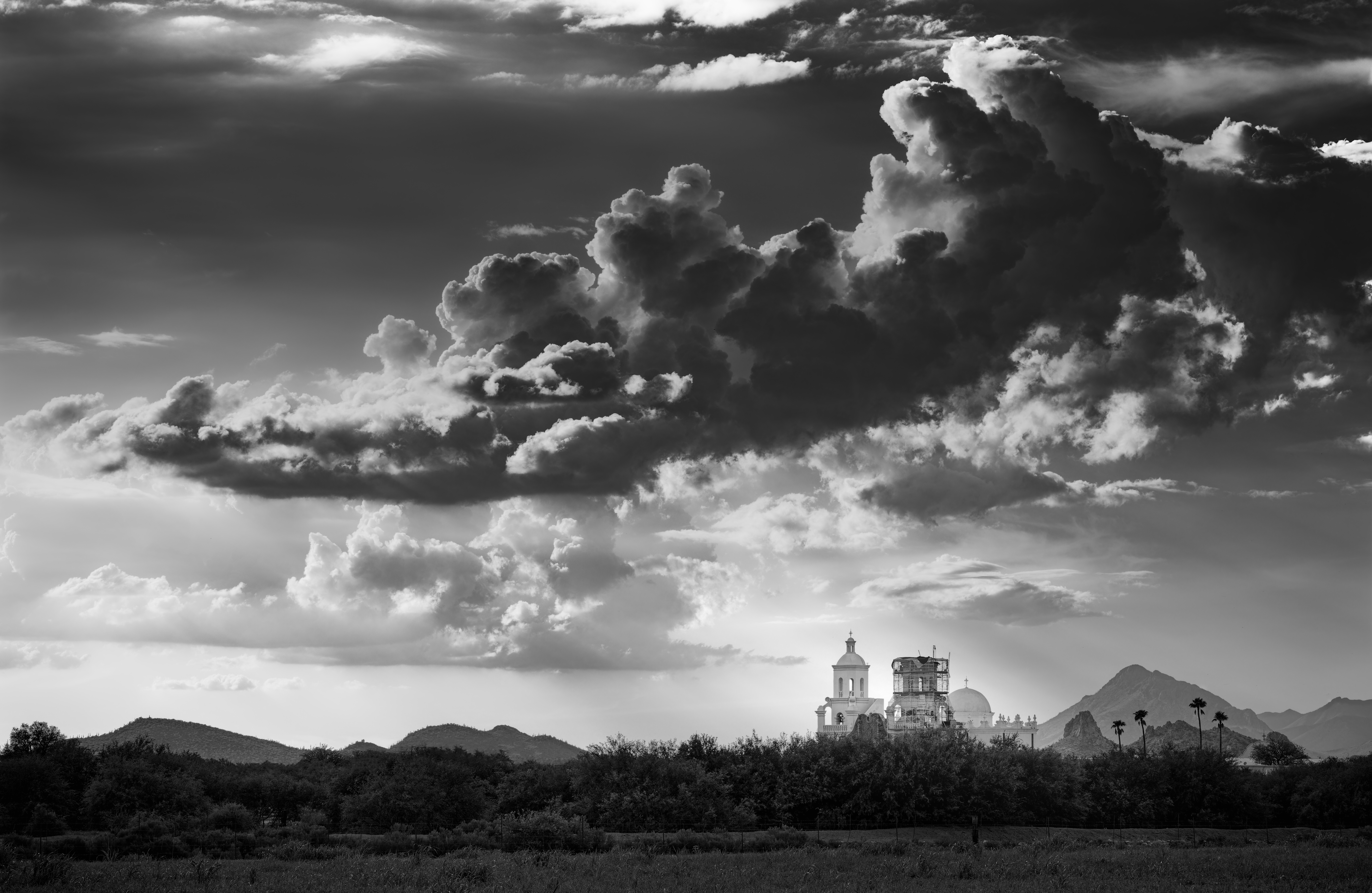 The Mission of San Xavier in black and white under billowing clouds with god-like rays shinning on the white mission. | Photo Title: La Misión de San Xavier del Bac | Photo by Jason Robert O'Kennedy ©2021