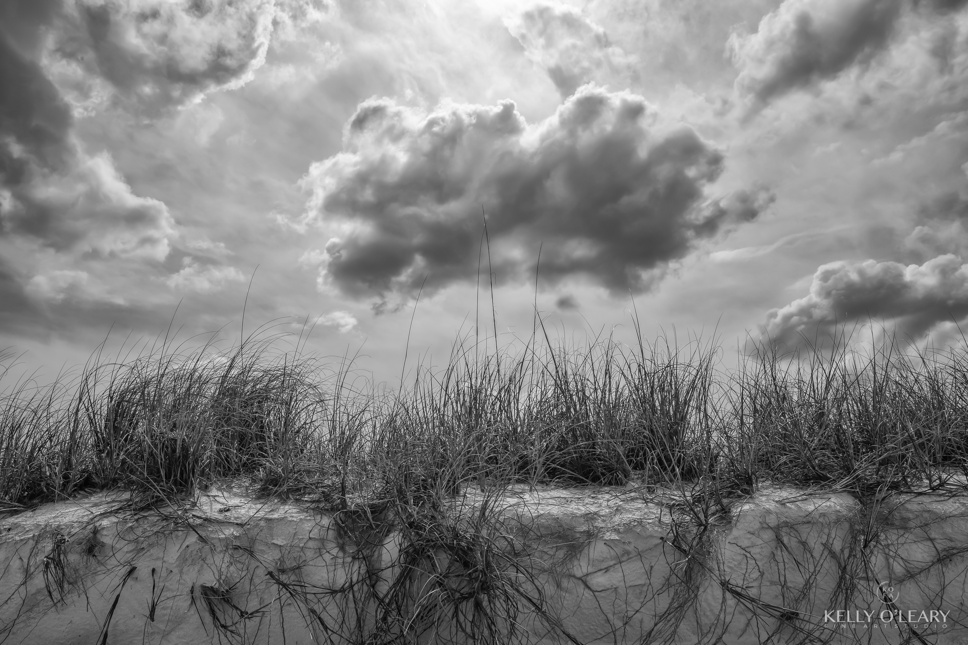A black and white image titled Stage Lighting photographed by Kelly O'Leary of a backlit cloud over a grassy beach bank. | Photo Title: Stage Lighting | 
        Photo by  ©2022